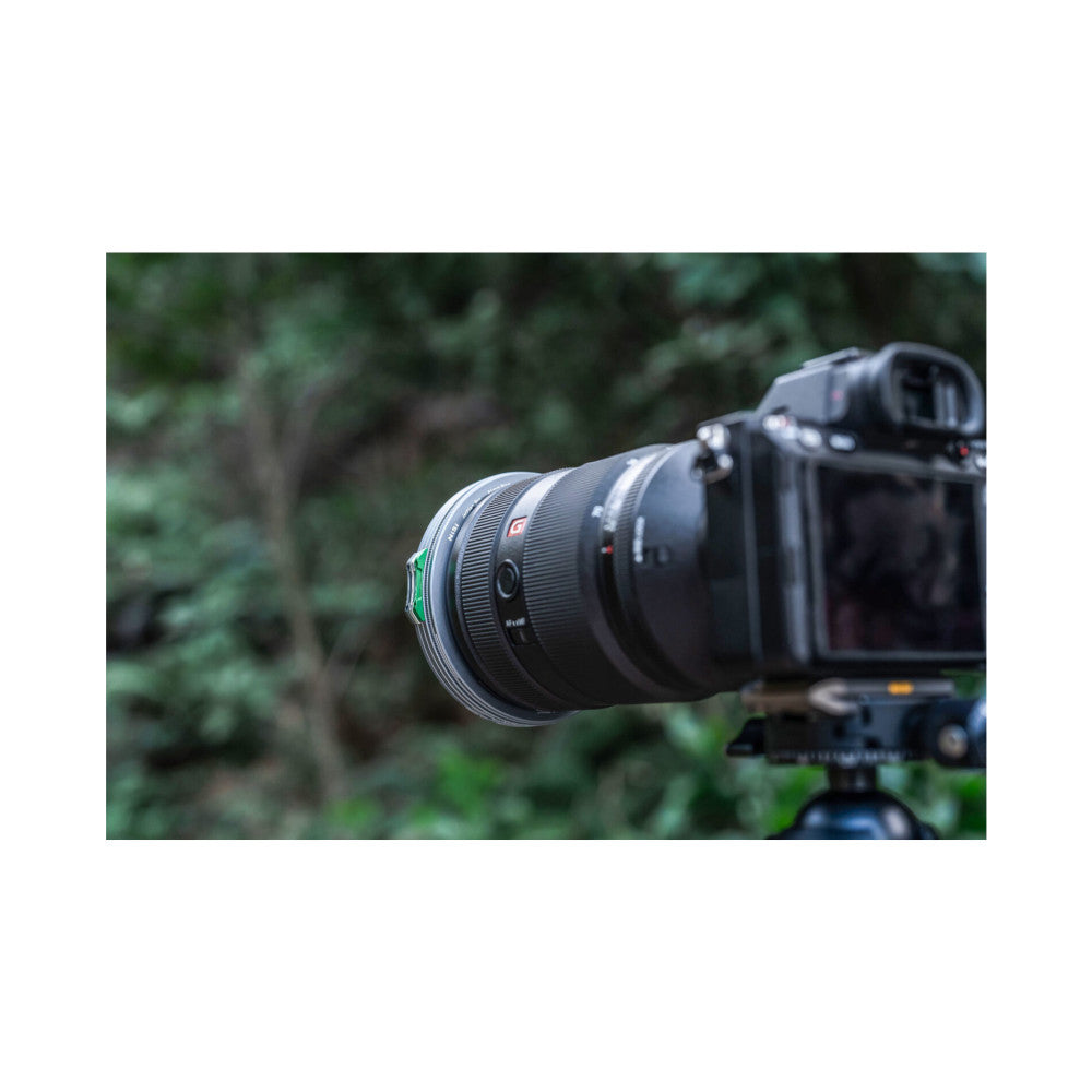 Professional camera with telephoto lens mounted on tripod, showing lens front element in close-up detail against blurred forest background.