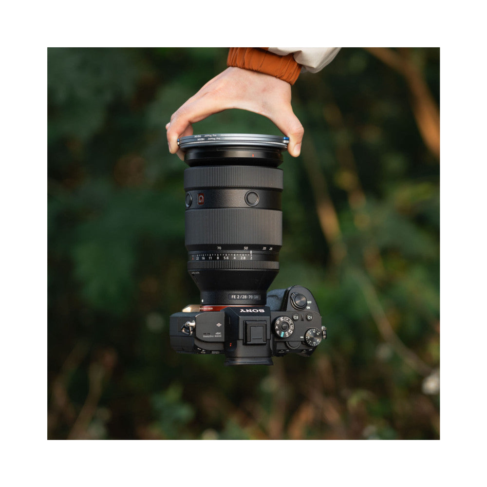 Hand holding Sony camera with 28-70mm lens, displaying NiSi magnetic filter ring attachment on lens barrel against blurred green background