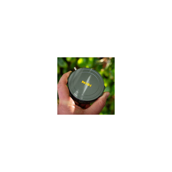 Hand holding a dark gray circular magnetic lens cap with yellow NiSi branding, photographed outdoors against a blurred green background.