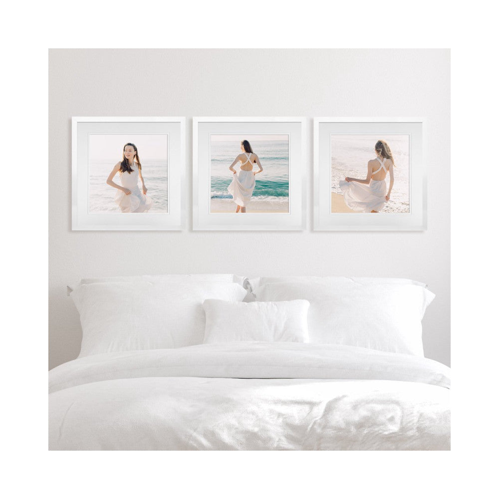 White bed with three framed beach portraits hung above headboard in minimalist bedroom setting.