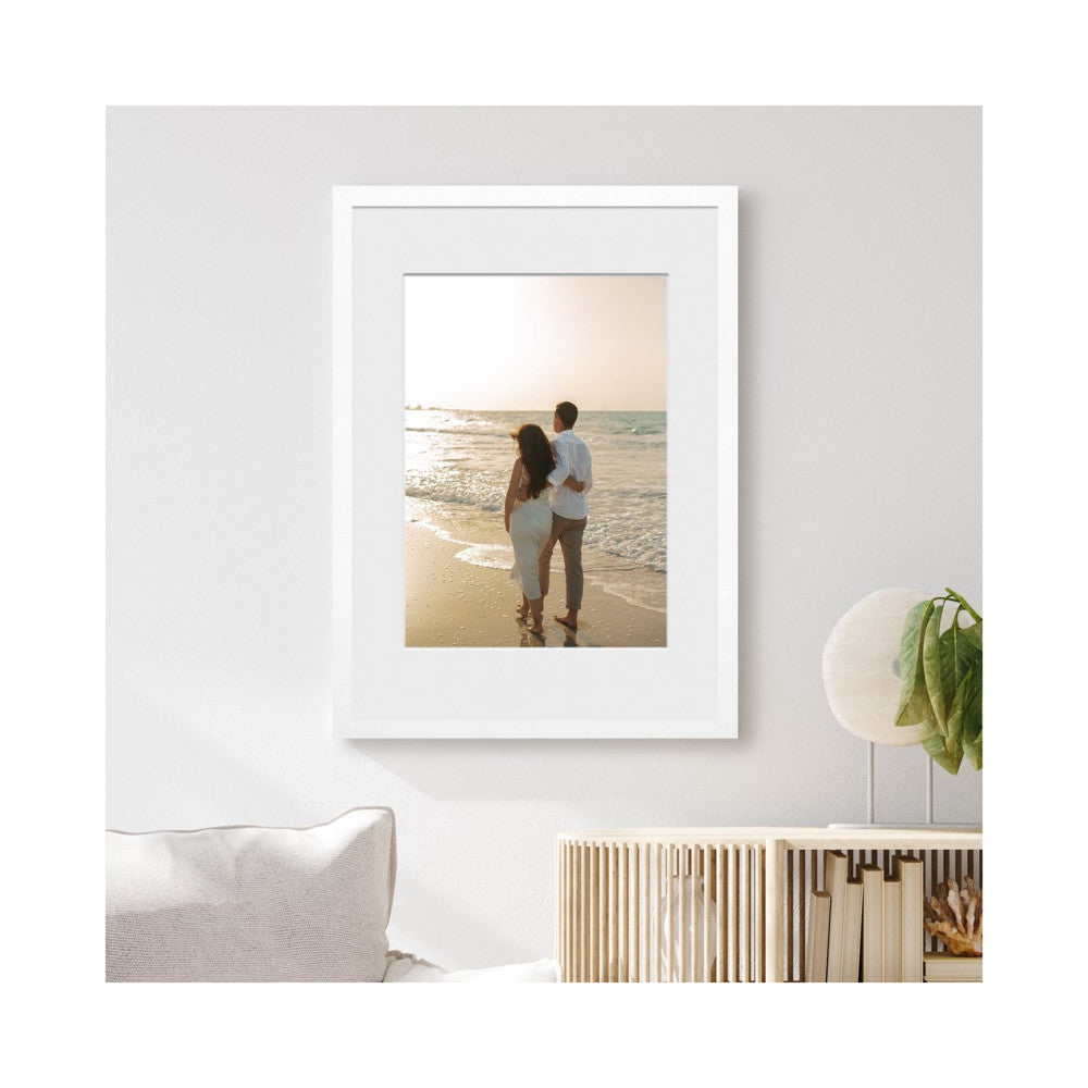 White framed photo display mounted on a light wall, showing a couple standing on a beach at sunset.