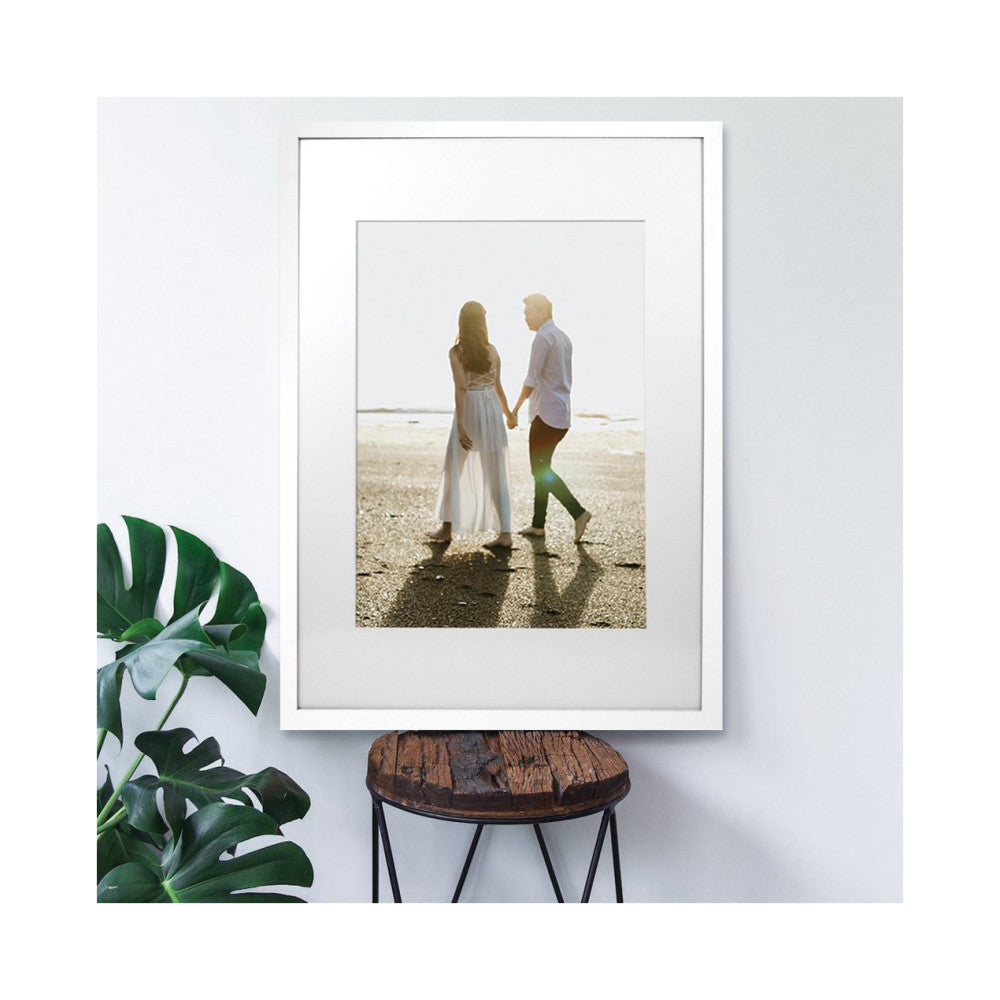White picture frame displaying a beach photograph of a couple holding hands, shown as a front-facing product display with a potted plant beside it.