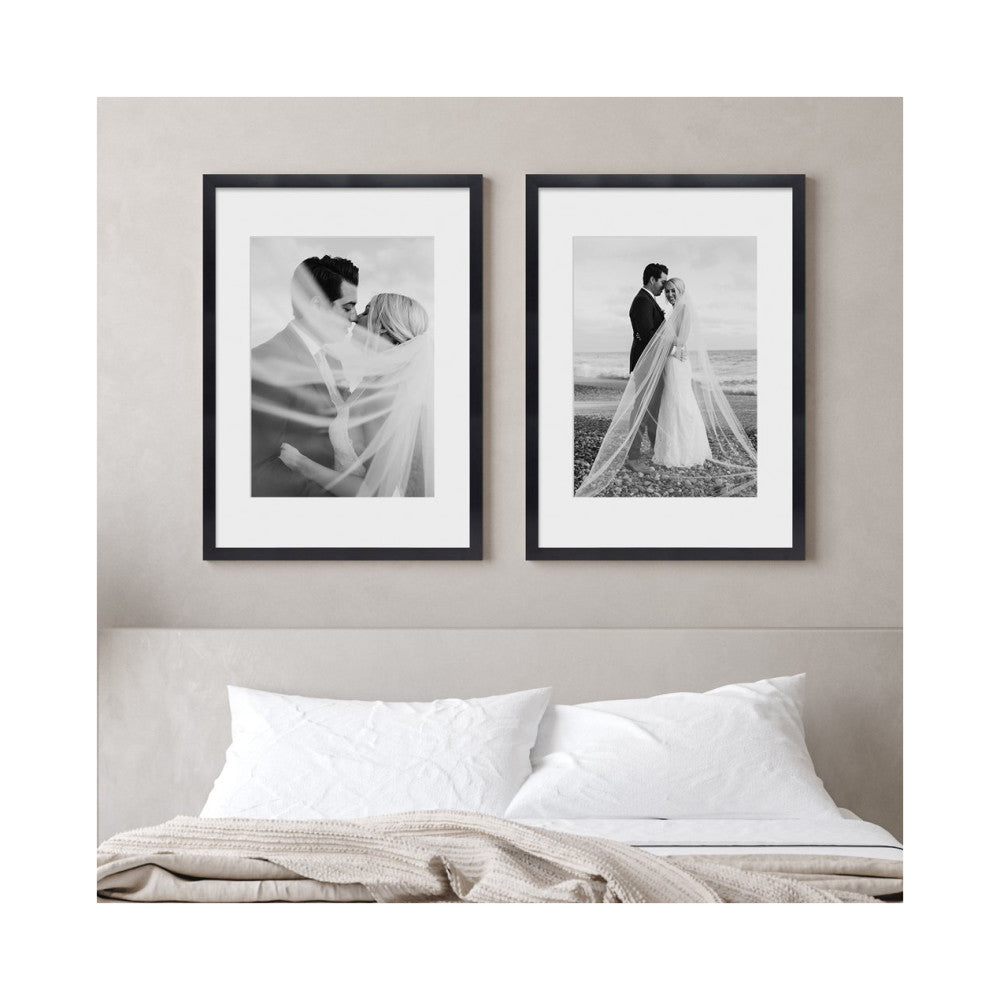 Two black framed wedding photographs displayed above a bed with white pillows and beige bedding against a neutral wall.