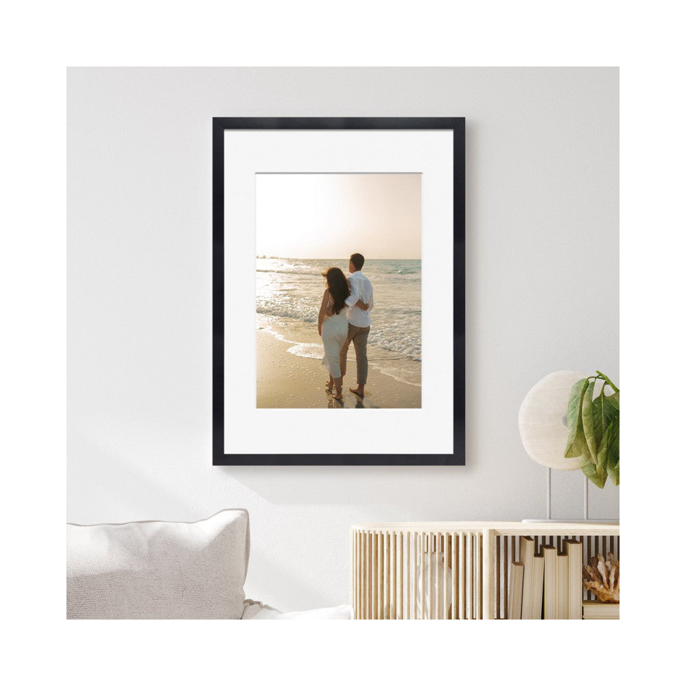 Black framed photo display mounted on white wall showing couple on beach at sunset, with white mat and cream cushion visible below.