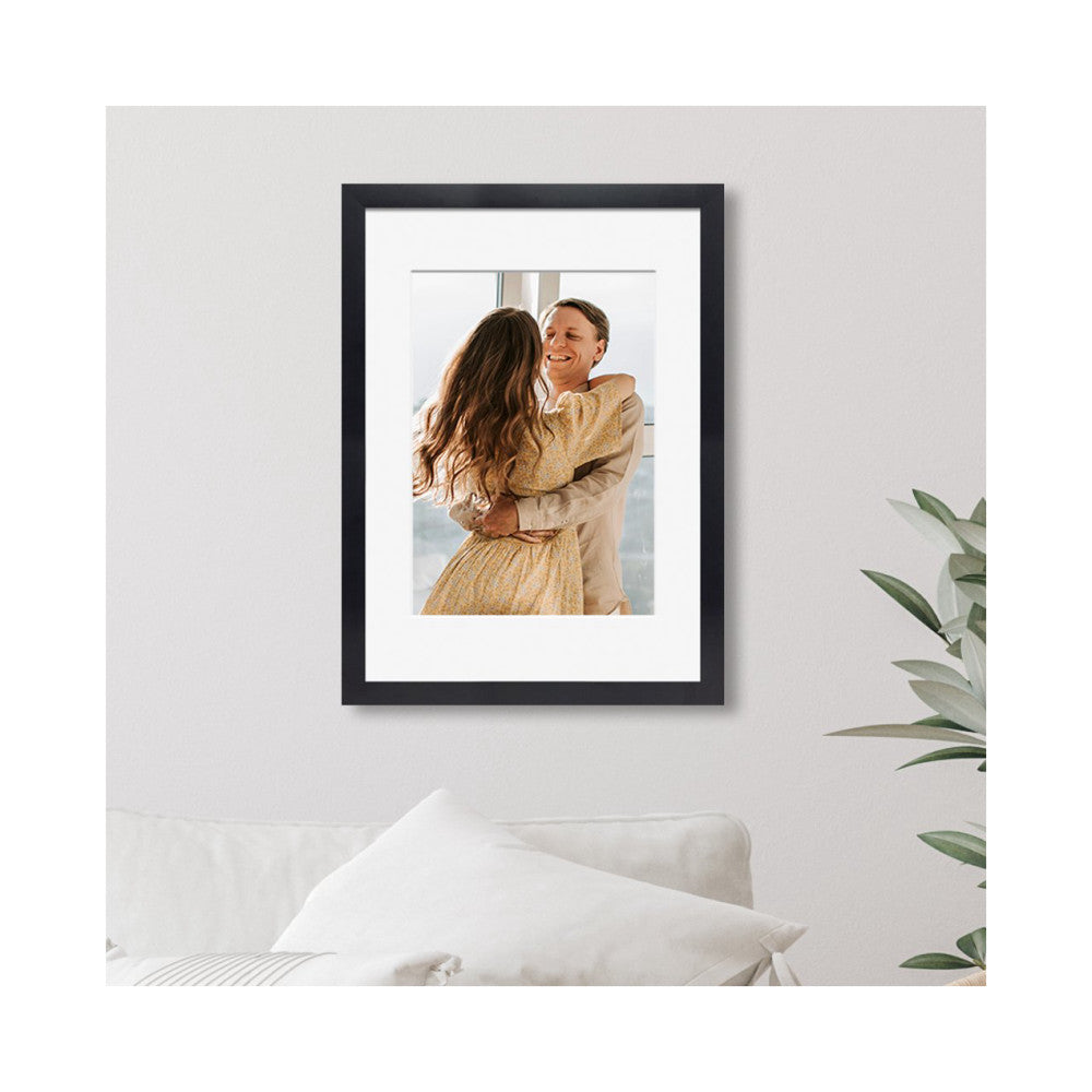 Black wooden frame displaying a portrait photo of a couple in warm-toned clothing, hung on a white wall above a white sofa with green plant visible on the right.