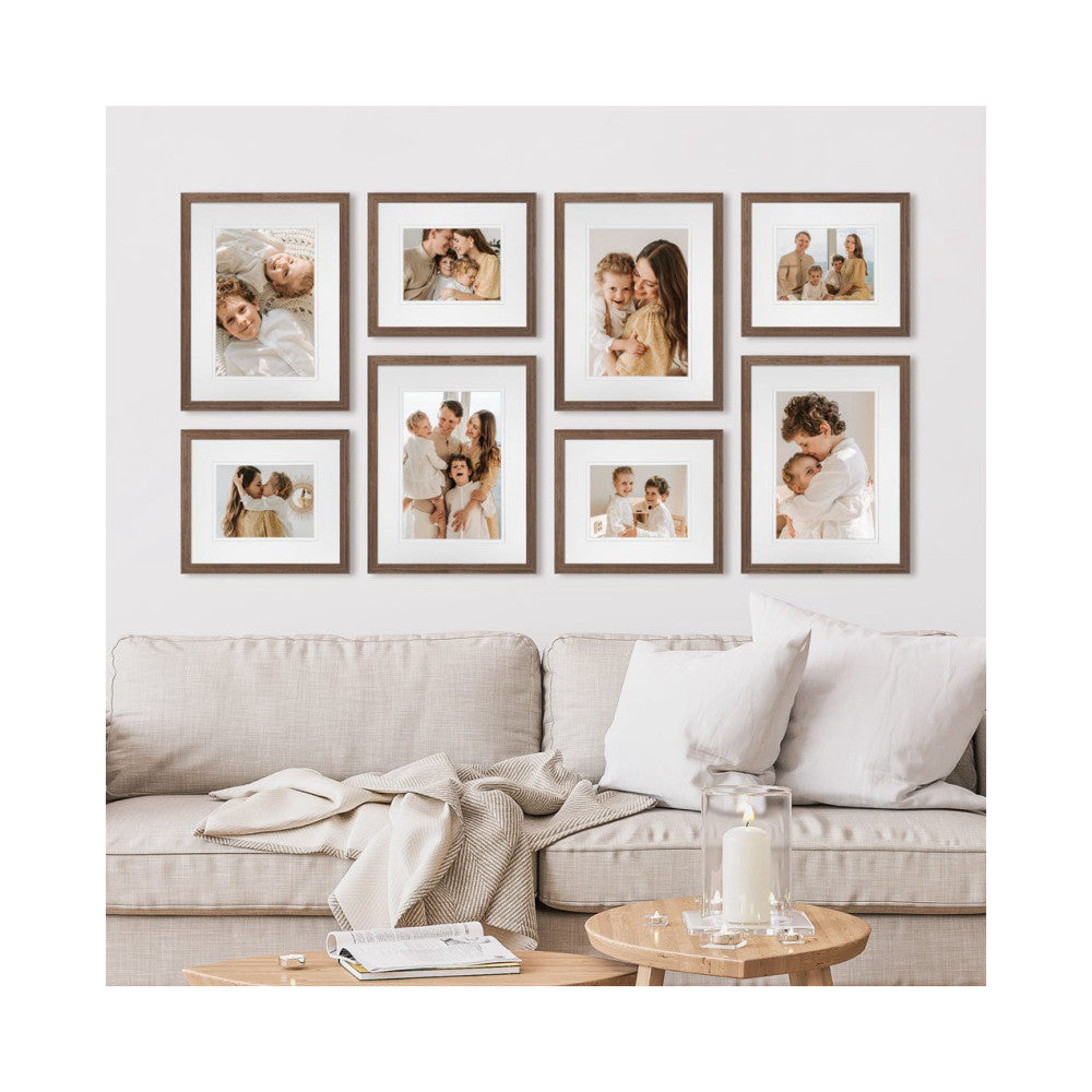 Eight brown wooden frames displaying family photos arranged on a light gray wall above a beige sofa with pillows and throws.