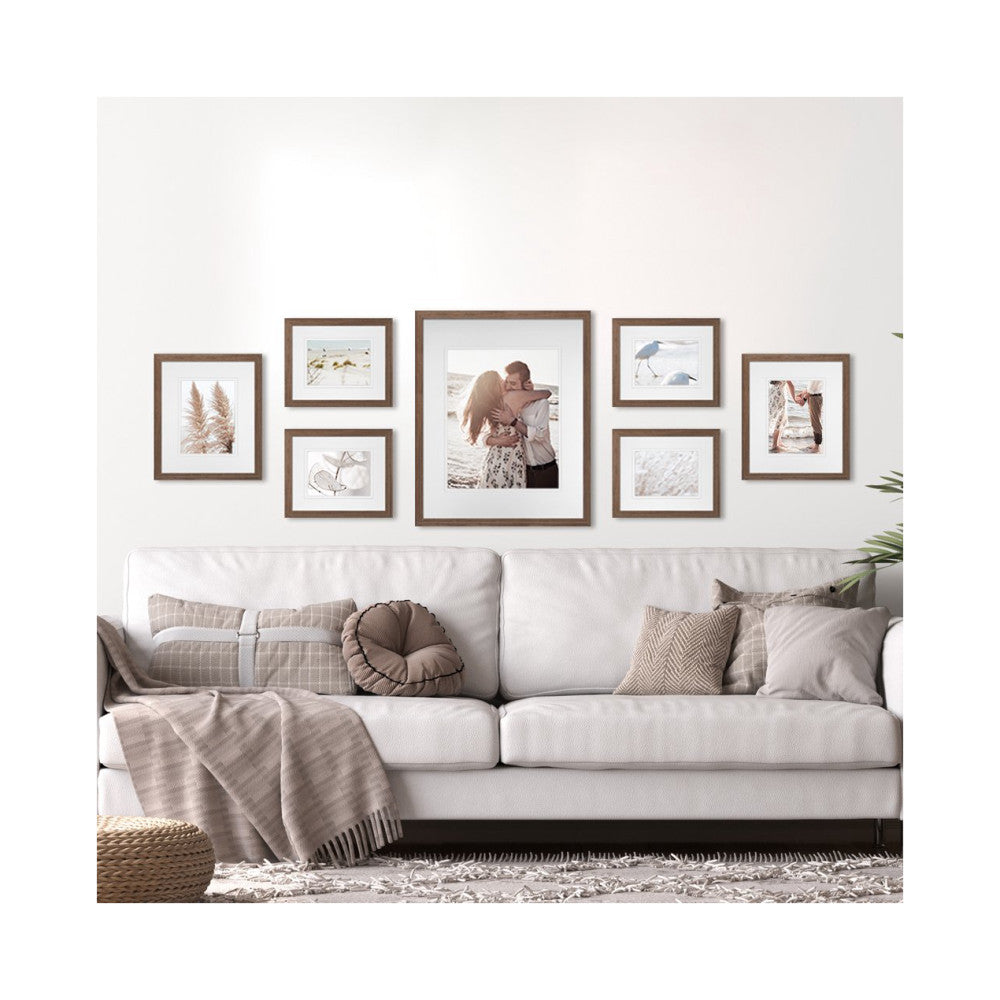 Gallery wall display above a cream sofa showing seven brown frames with white mats containing photos and artwork in a living room setting.