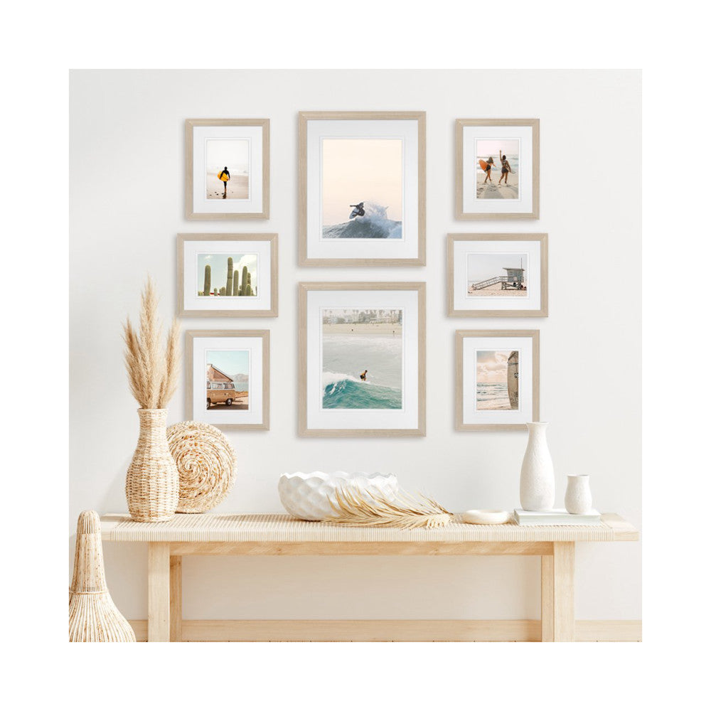 Gallery wall with eight framed beach and nature photos in light wood frames arranged on white wall above wooden console table with decorative accessories.