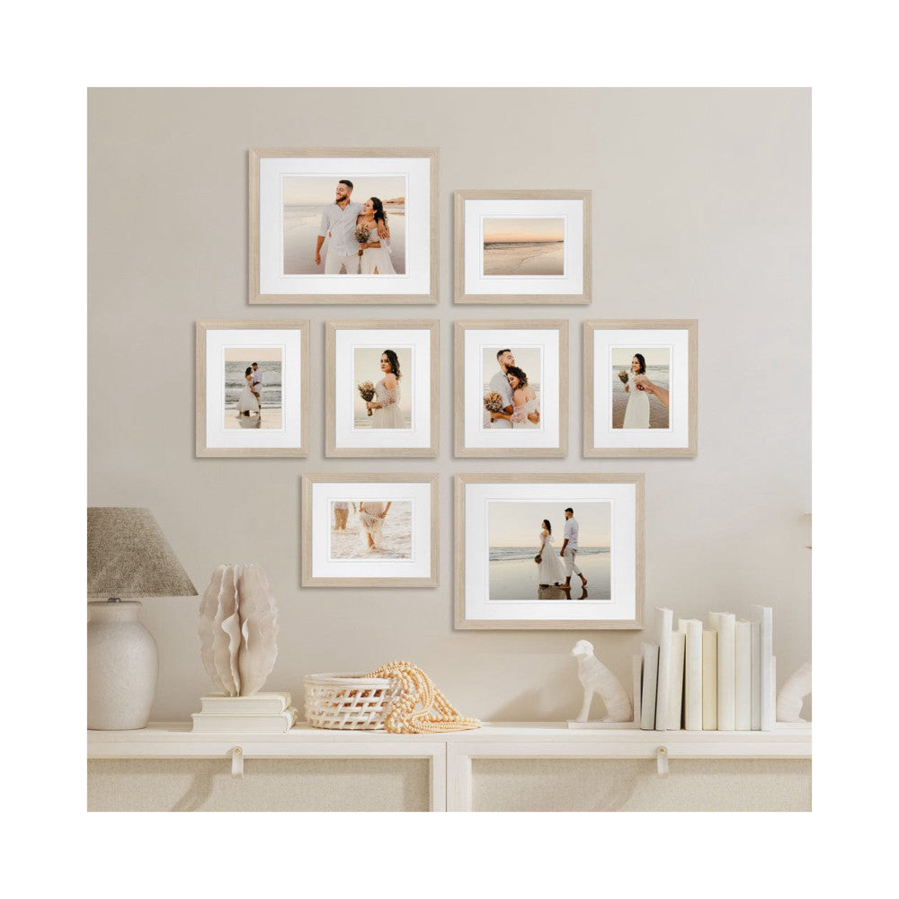 Eight wooden frames with white mats displaying beach wedding photos arranged on a beige wall above a white shelf with home decor.