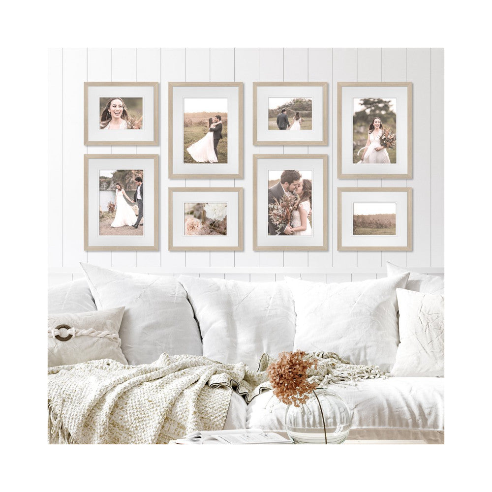 Eight wooden frames with white mats displaying wedding photos arranged on a white shiplap wall above a white sofa.