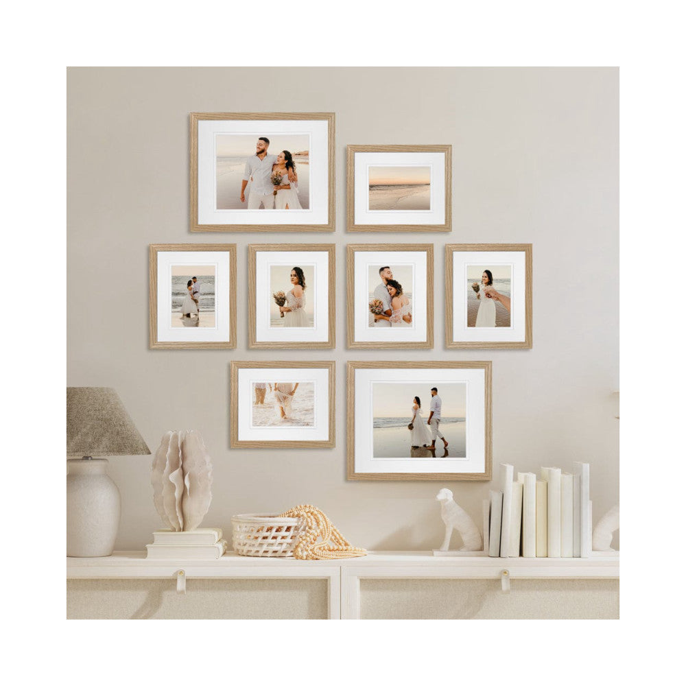 Eight natural oak frames displaying beach wedding photos arranged on a beige wall above a white shelf with decorative objects.