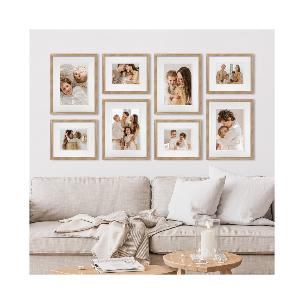 Eight natural oak-framed family photos arranged on a white wall above a beige linen sofa with neutral decor and a wooden table.