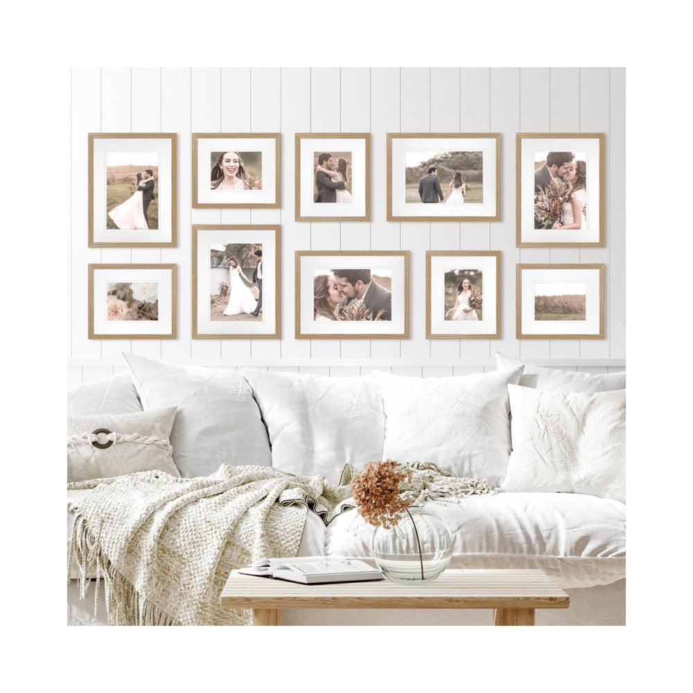 In-use gallery wall display showing ten wooden frames with wedding photos arranged above a white sofa in a bright, minimalist bedroom setting.