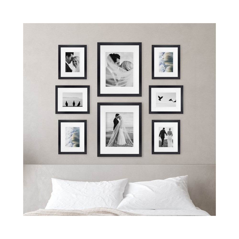 Eight black-framed photos arranged on a bedroom wall above a bed with white pillows, displaying wedding and nature photography.