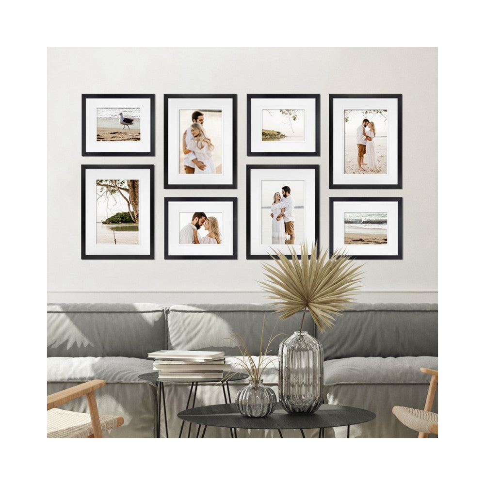 Eight black framed photos arranged on a wall above a gray sofa, displaying beach and couple portrait images in a living room setting.