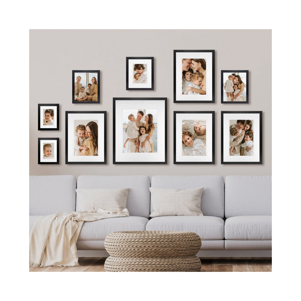 Ten black framed family photos arranged on a gray wall above a light gray sofa with decorative pillows and a woven pouf.