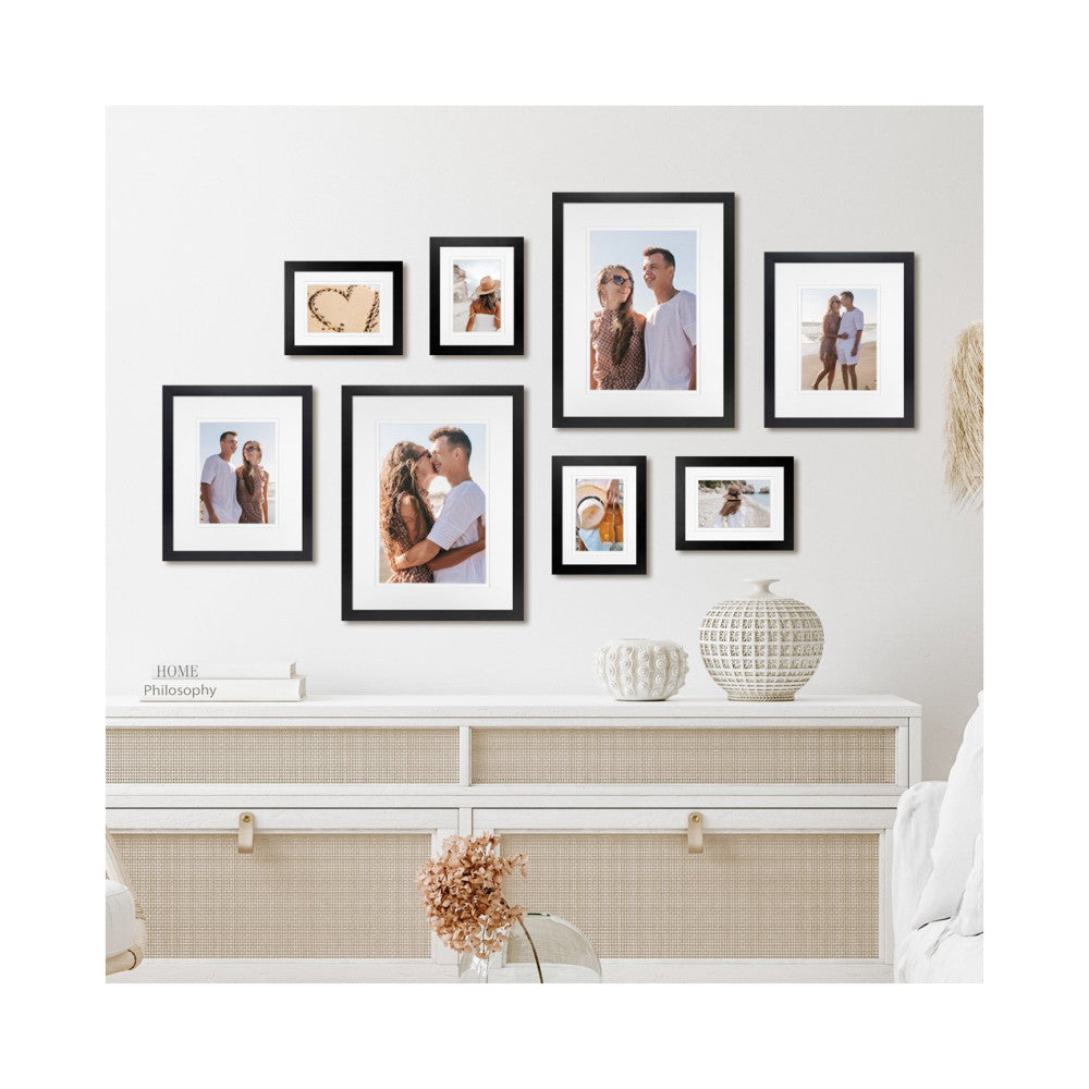 Eight black frames arranged on white wall above dresser displaying beach family photos and decorative items.