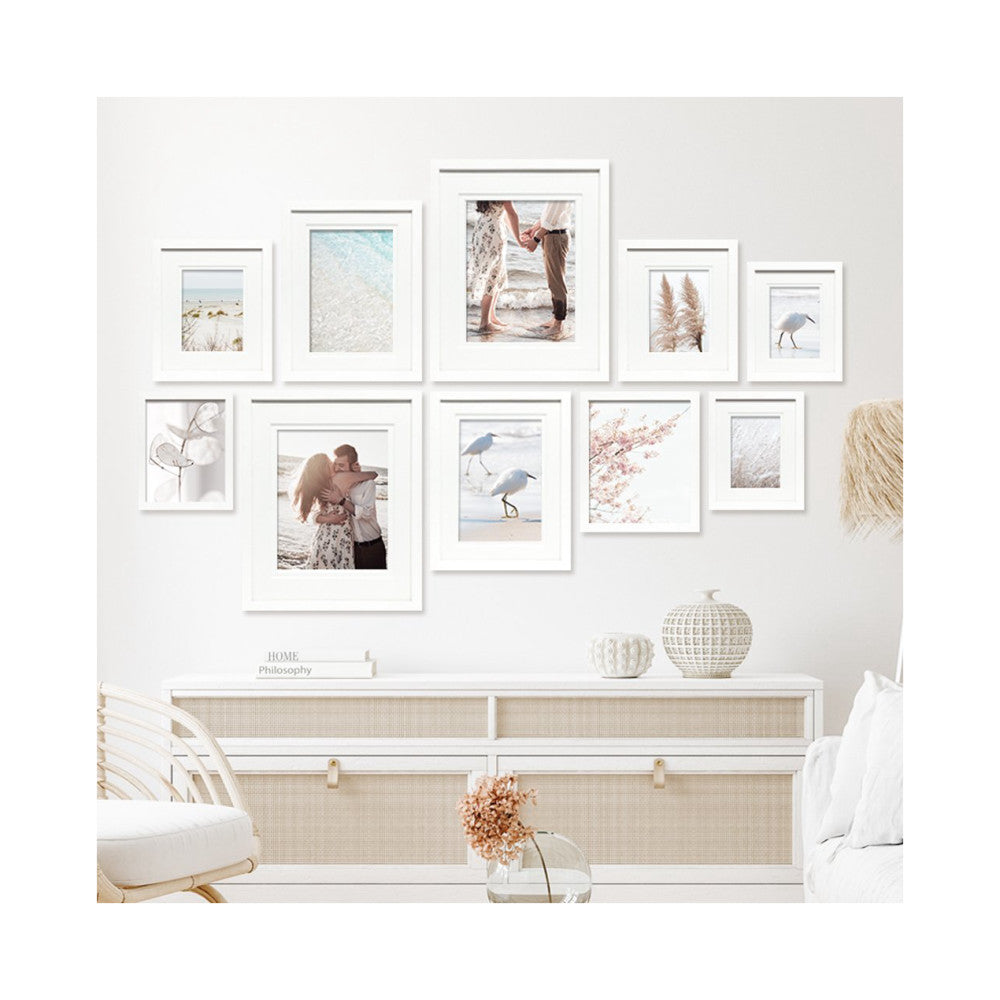 White framed gallery wall display with 10 frames arranged in an asymmetrical pattern above a light wood dresser in a coastal-themed bedroom interior.