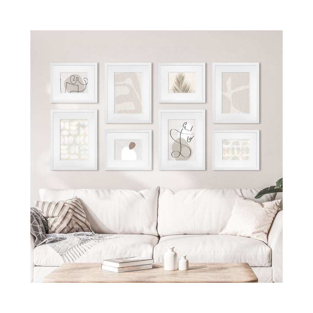 Eight white framed artworks arranged on a beige wall above a cream sofa with decorative pillows and a wooden table.