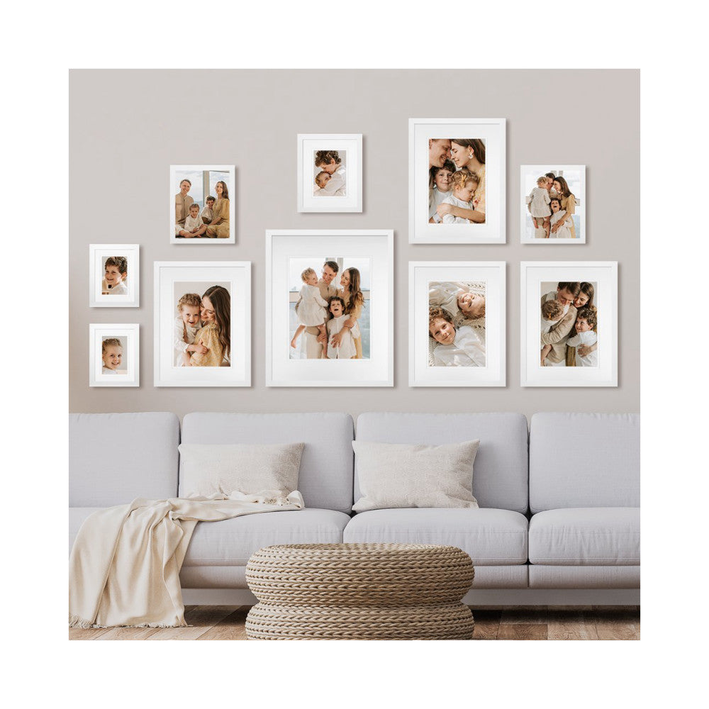 White framed family photos arranged on a beige wall above a gray sofa with decorative pillows and woven ottoman.
