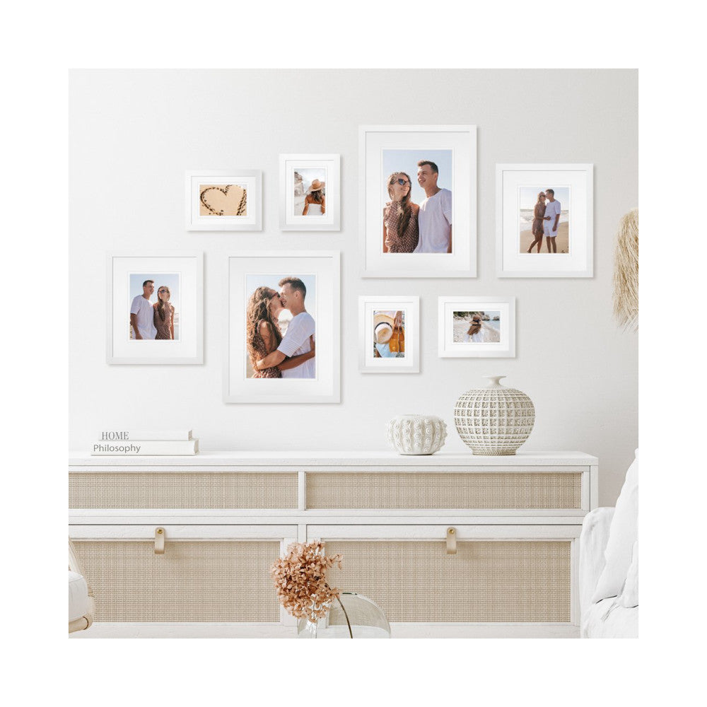 Eight white framed photos arranged on a light wall above a white dresser with decorative items and beach-themed artwork.